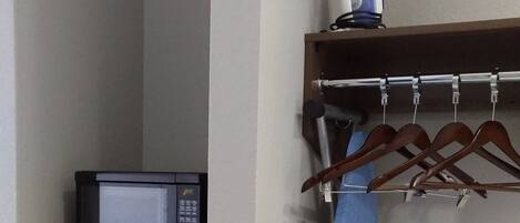 Una kitchenette compacta con microondas y refrigerador, una toalla azul colgada en un perchero, y una tabla de planchar fijada a la pared.
