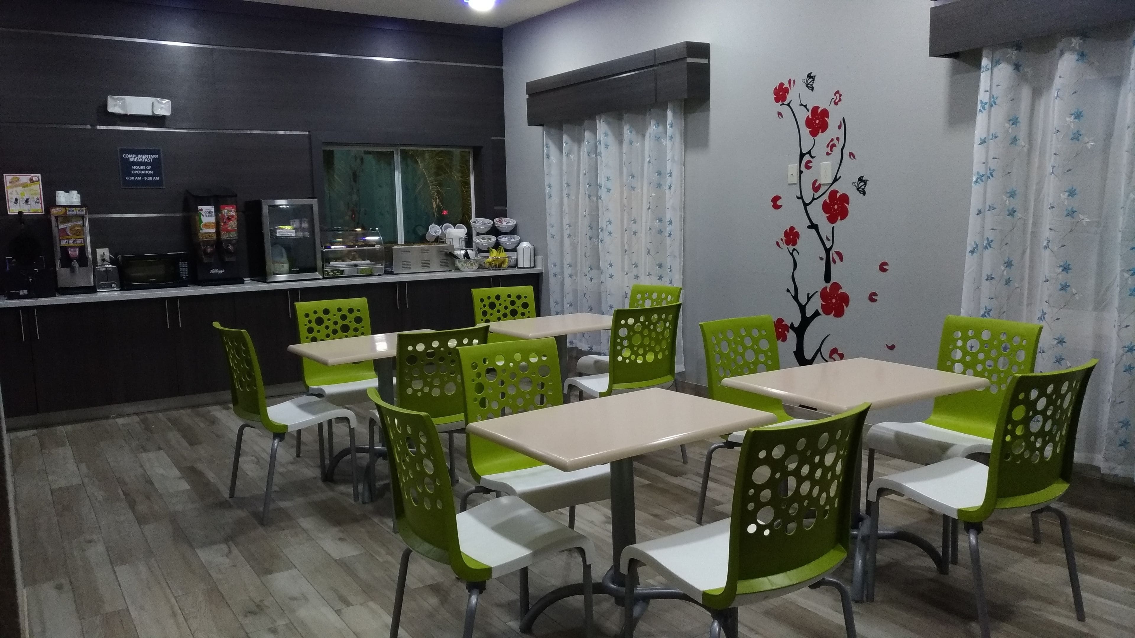 A dining area with green chairs and tables, a kitchen area with a refrigerator and microwave, and a wall with a floral mural.