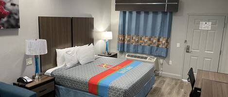 Habitación de hotel con cama, mesitas de noche, escritorio, silla, lámpara, teléfono, calefactor y puerta.