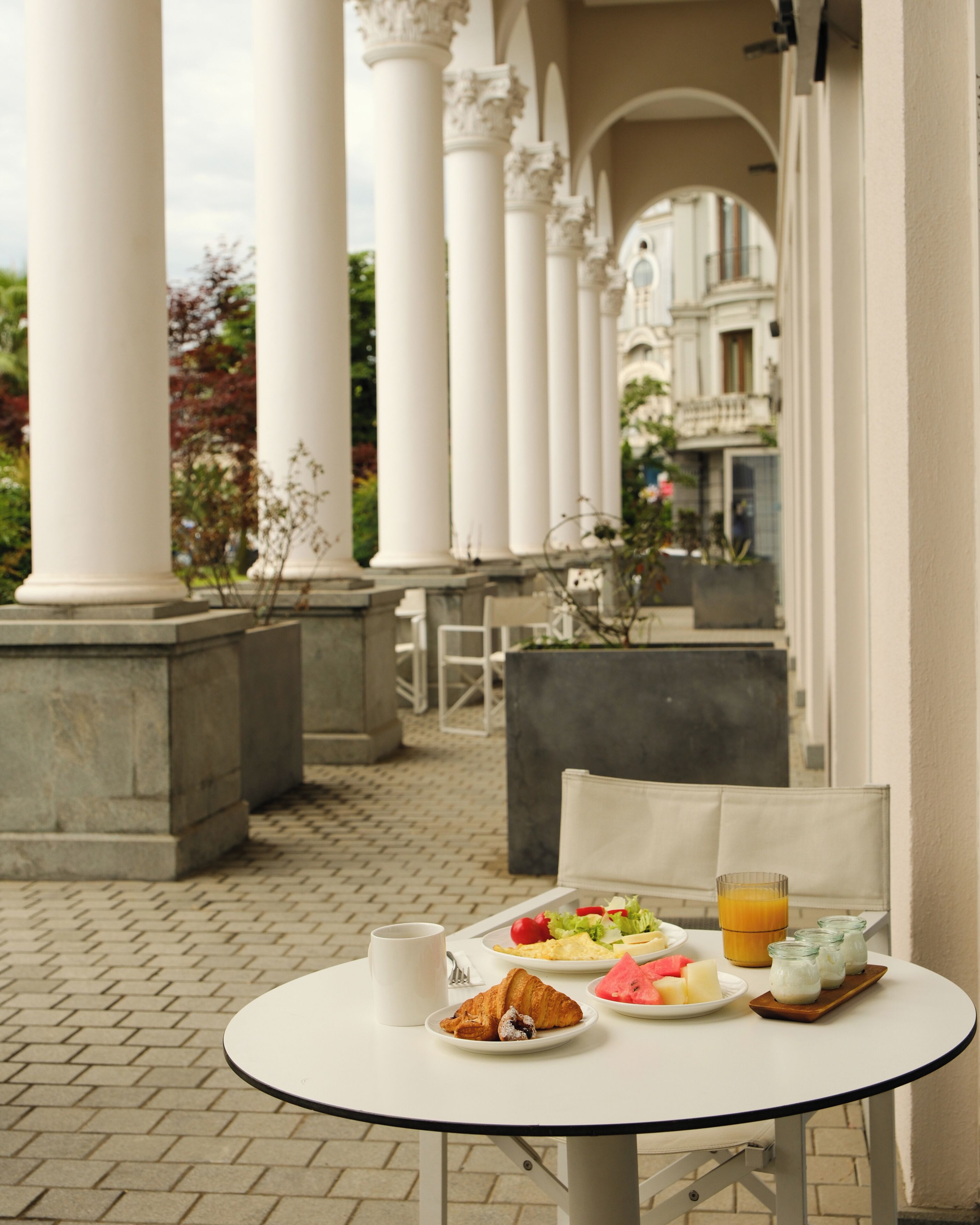 A table with breakfast items, including croissants, fruit, and juice, in a courtyard with columns and a view of buildings.