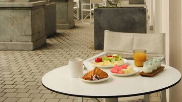 A table with breakfast items, including croissants, fruit, and juice, in a courtyard with columns and a view of buildings.