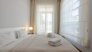 A hotel room with a large bed, a nightstand with a lamp, and a view of the city through sheer curtains. - Le Port Apart Hotel (Batumi)