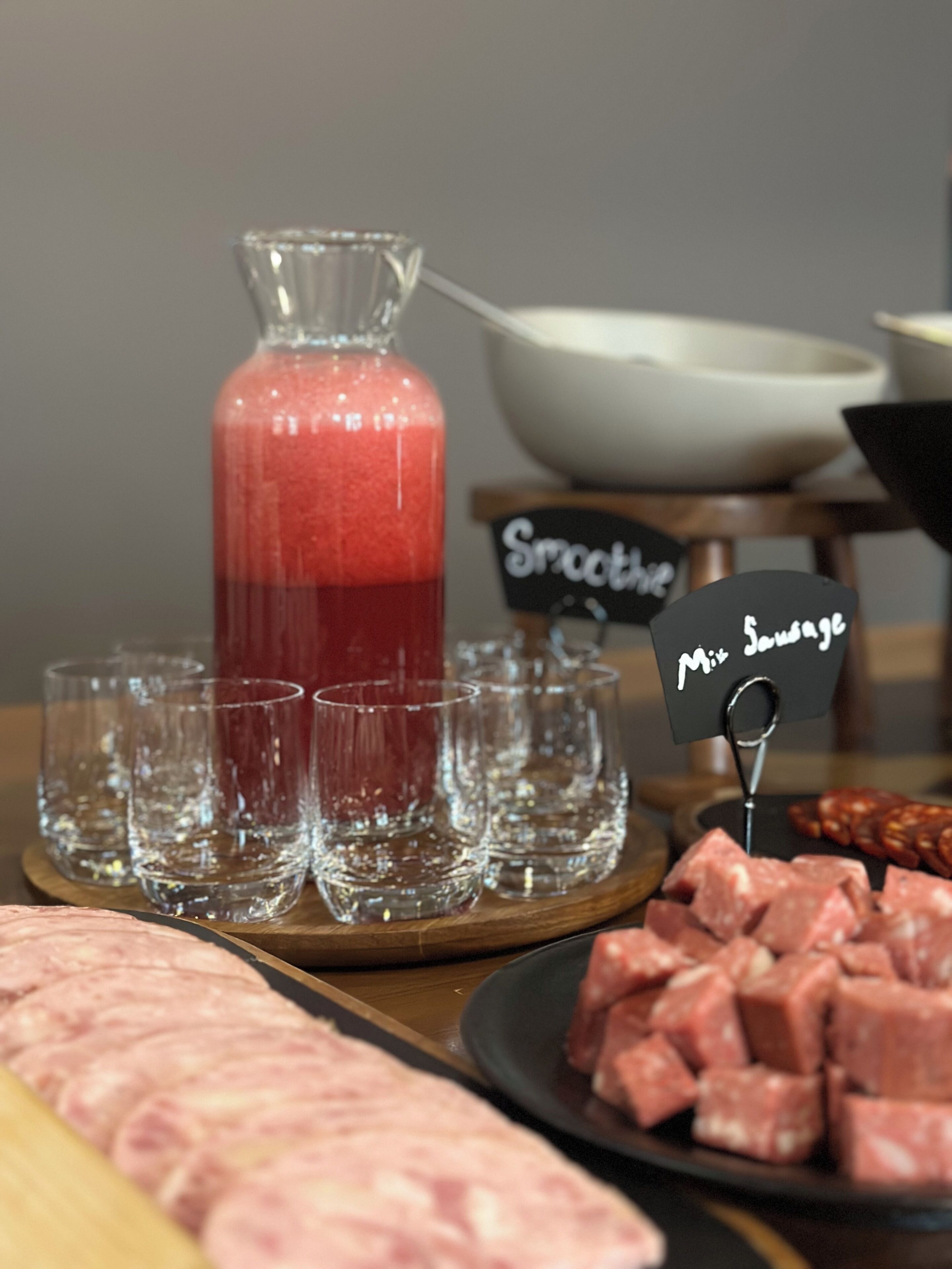 En buffetbod med en kande rød smoothie, flere glas og en tallerken med skåret kød.