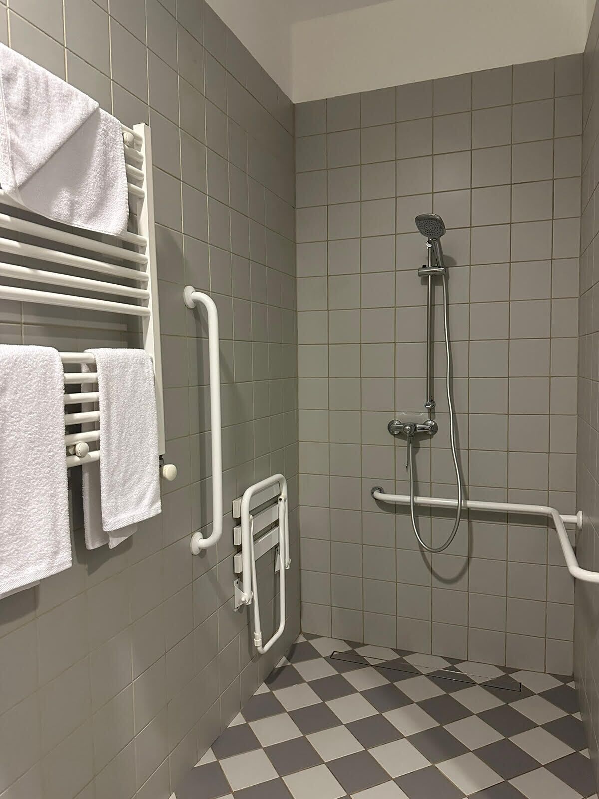 A bathroom with grey tiled walls, a checkered floor, a white towel rack, a shower with a handheld showerhead, and a grab bar.