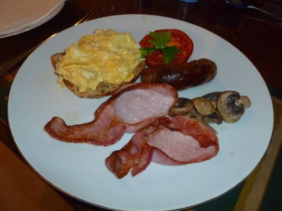 A plate with scrambled eggs, sausages, bacon, mushrooms, and a tomato on a white plate.