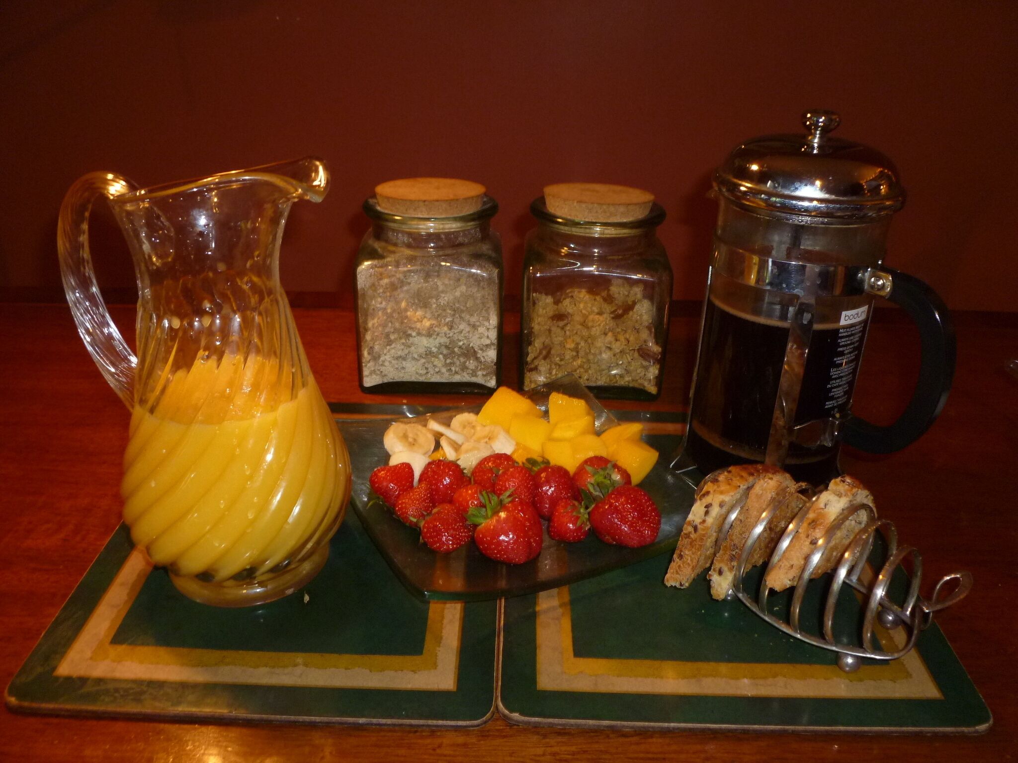 Desayuno servido con un jarro de jugo de naranja, una fuente de fruta, pan rebanado y dos recipientes de comida.