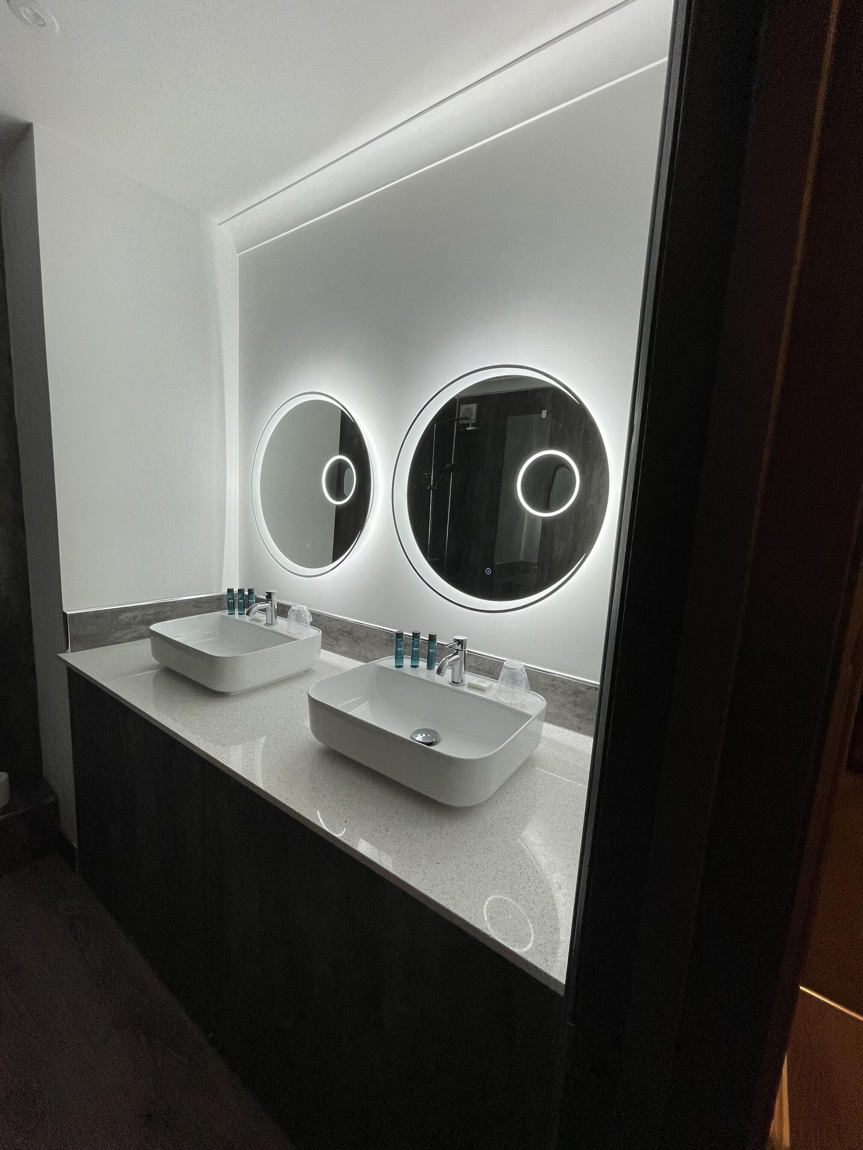 Un baño moderno con dos lavamanos, un espejo grande y luces circulares sobre los espejos.