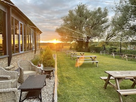 A patio area with outdoor seating and picnic tables, a building with large windows, and a sunset in the background.