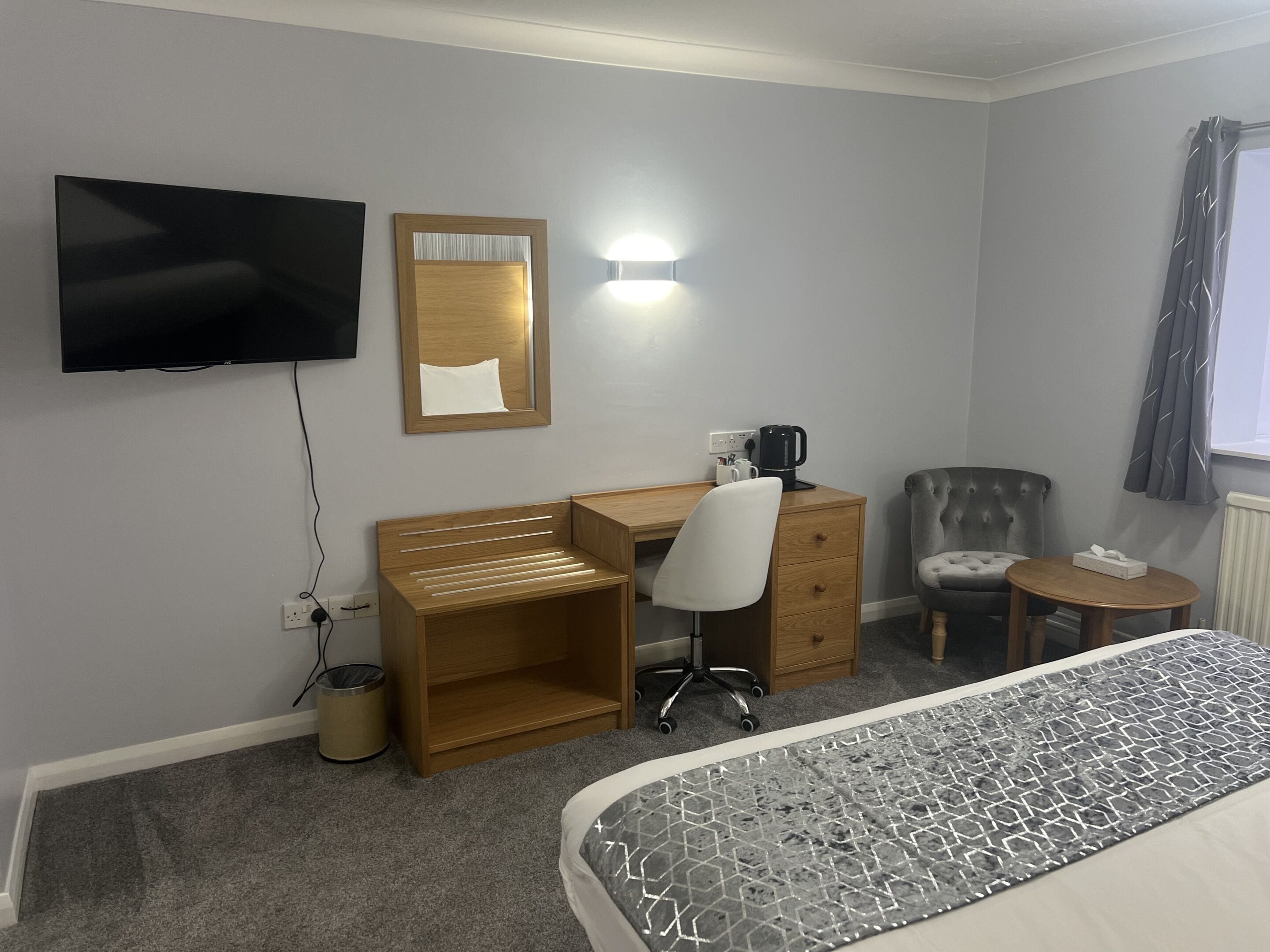 A hotel room with a bed, a desk, a chair, a television, a mirror, a lamp, and a window with curtains.