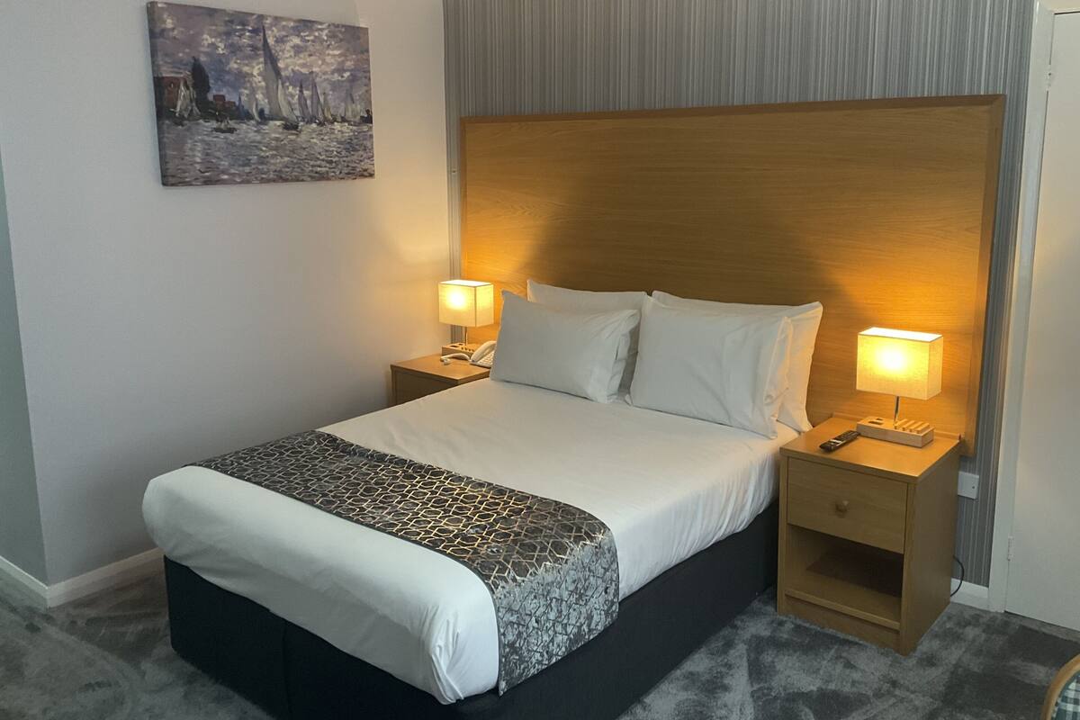 A hotel room with a large bed, two bedside tables with lamps, a wooden headboard, a painting on the wall, and a carpeted floor.