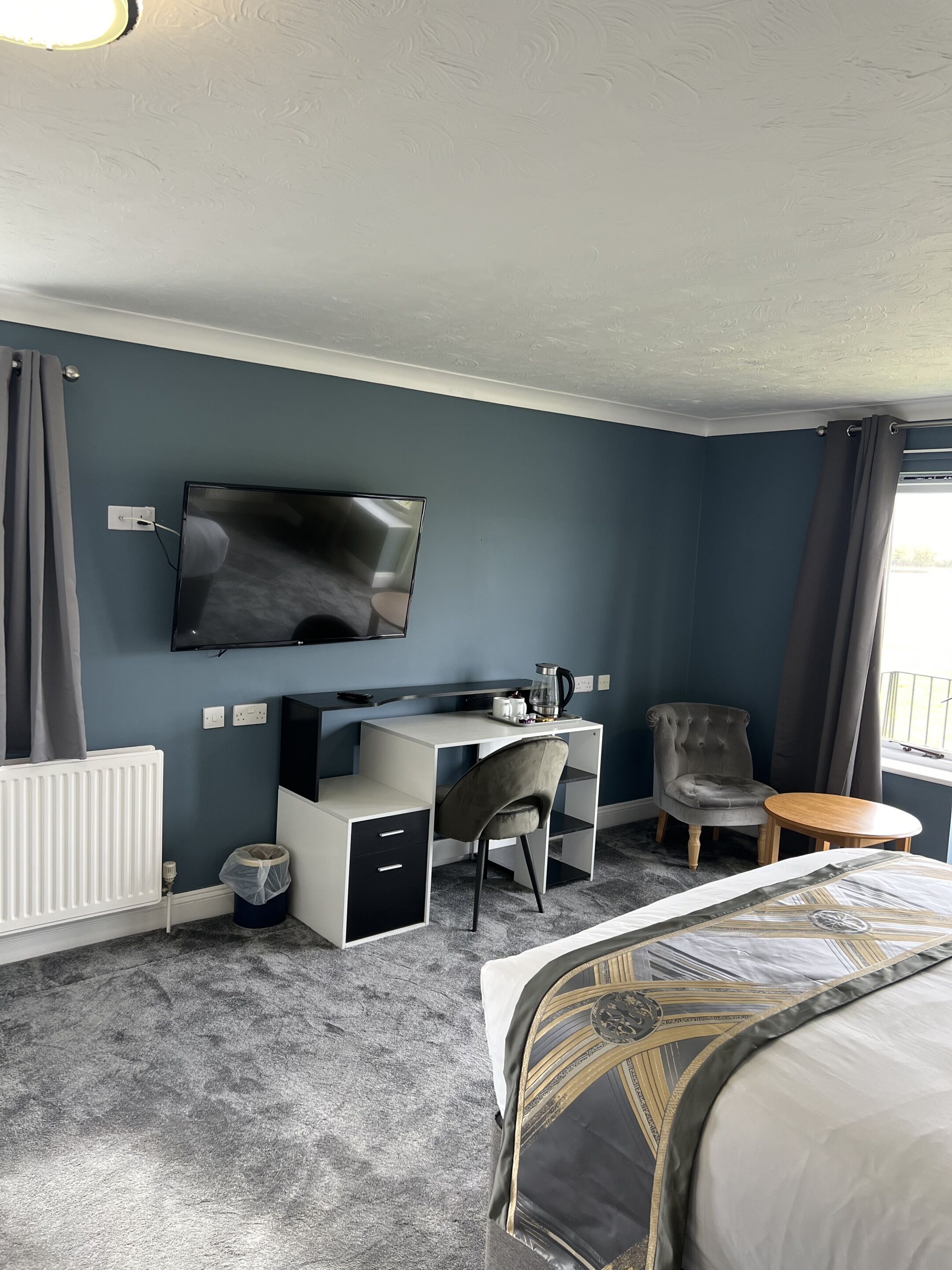 Habitación de hotel con cama, escritorio, silla, televisor, radiador y ventana con cortinas.