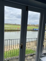 Vista desde el interior de una habitación con puerta a un balcón que da a un río, y una aguja de iglesia a lo lejos.