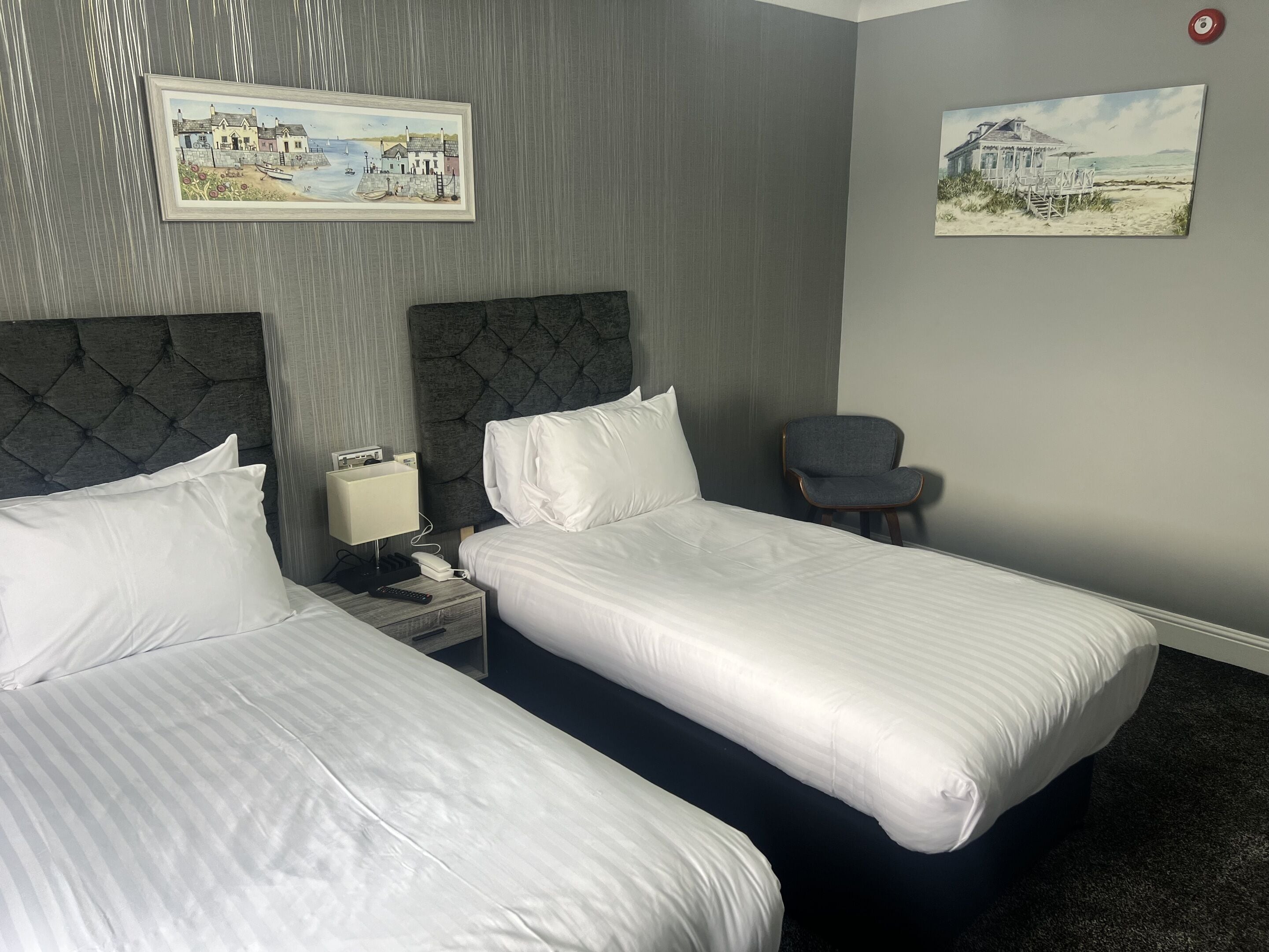 A hotel room with two beds, a chair, a nightstand with a lamp, and two paintings on the wall.