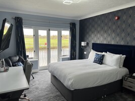 A hotel room with a large bed, a desk with a chair, a TV, and a view of a green landscape through the windows.