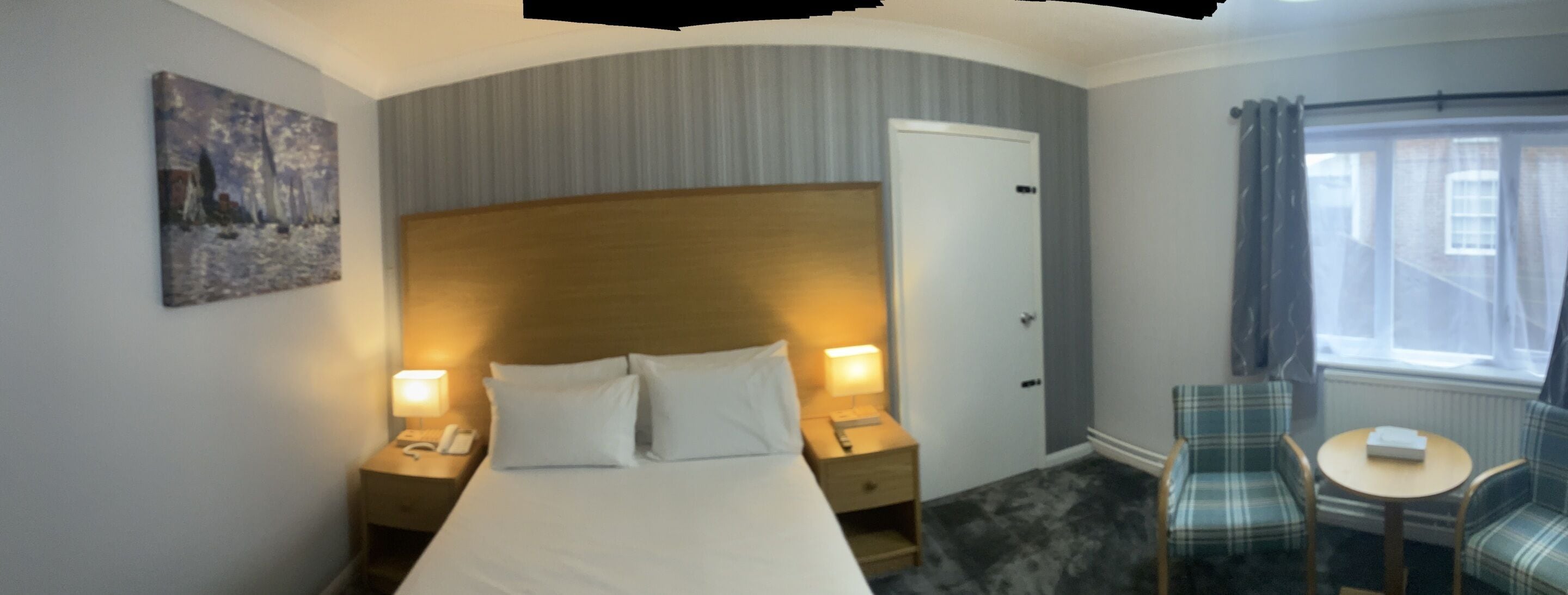 A hotel room with a bed, bedside lamps, a small table, a chair, and a painting on the wall.