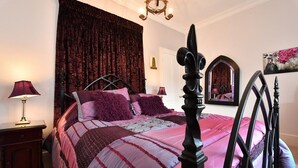 A bedroom with a large bed, dark curtains, a chandelier, a nightstand with a lamp, and a mirror on the wall. - Heritage Bed & Breakfast (Calne)