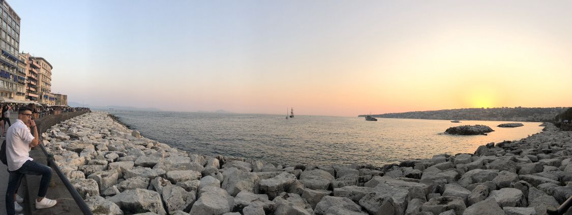 A coastal view with a rocky breakwater, a calm sea, and a sunset in the distance.