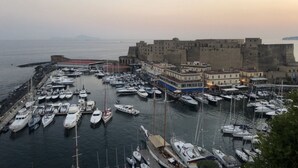 Un port de plaisance où de nombreux bateaux sont amarrés, un grand bâtiment qui ressemble à une forteresse, et une mer calme.