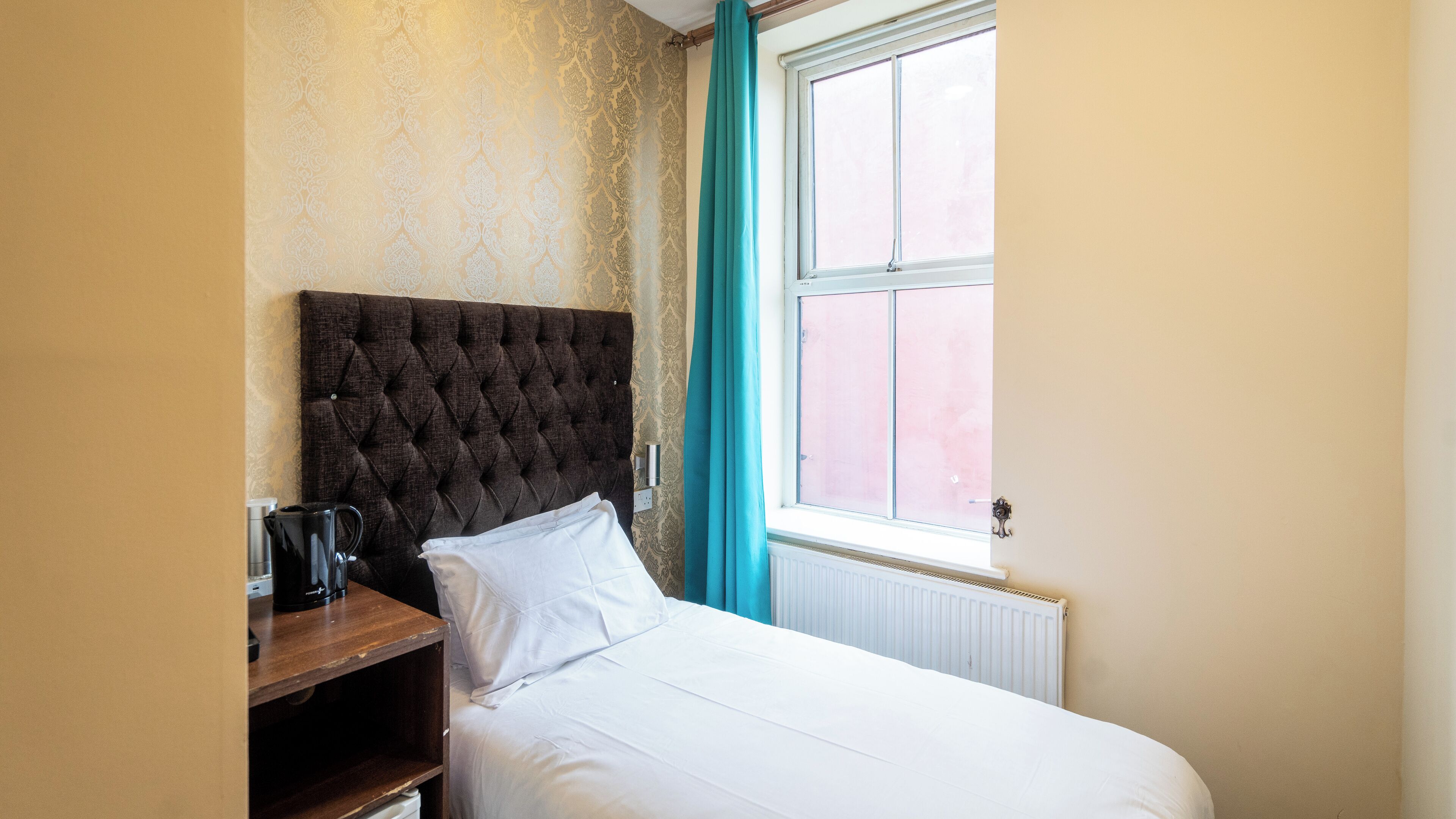A bedroom with a tufted headboard, a white bed, a wooden nightstand, a kettle, and a window with a view of a pink building.
