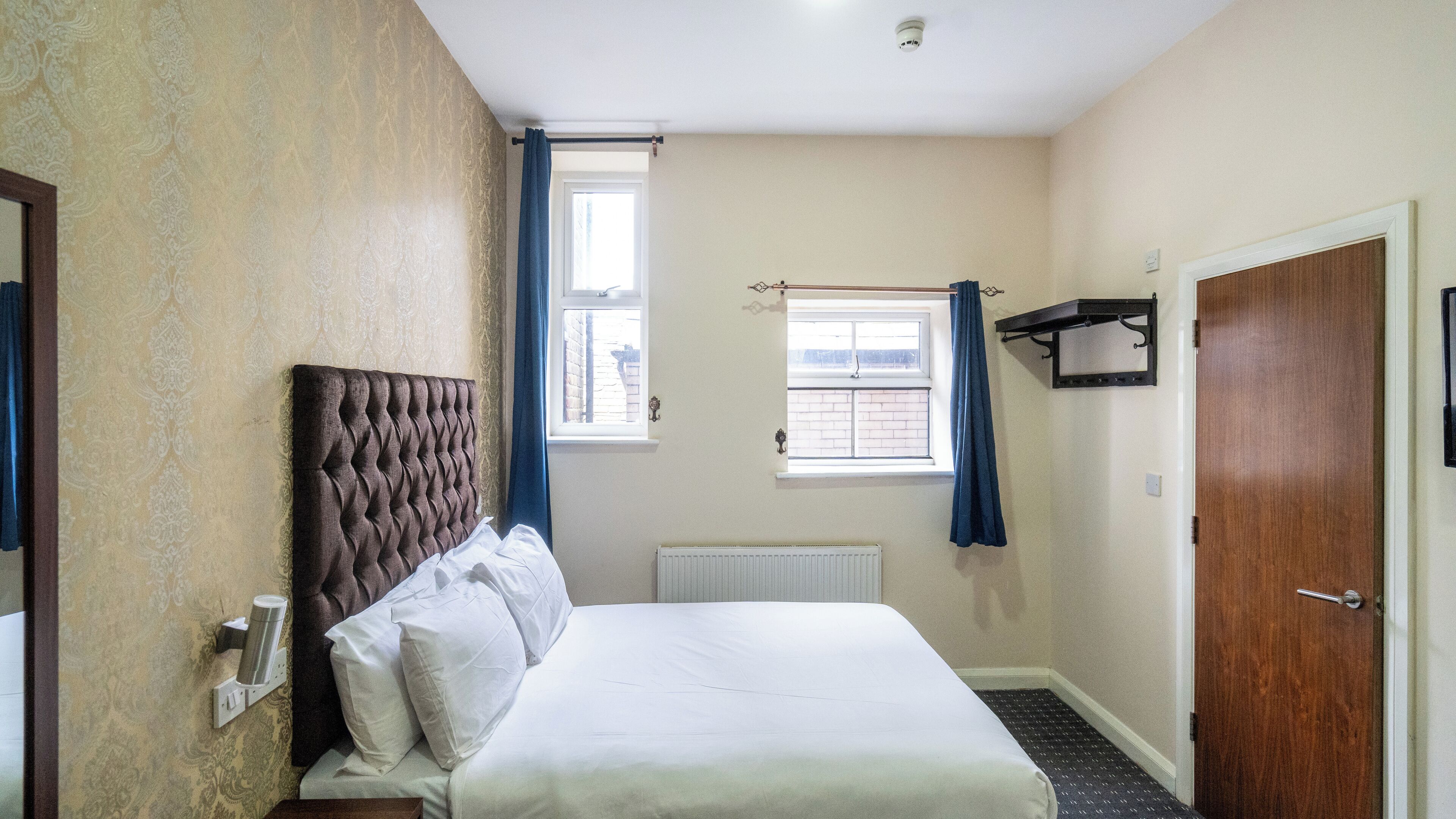 A hotel room with a large bed, a radiator, a window with curtains, a door, and a wall-mounted shelf.