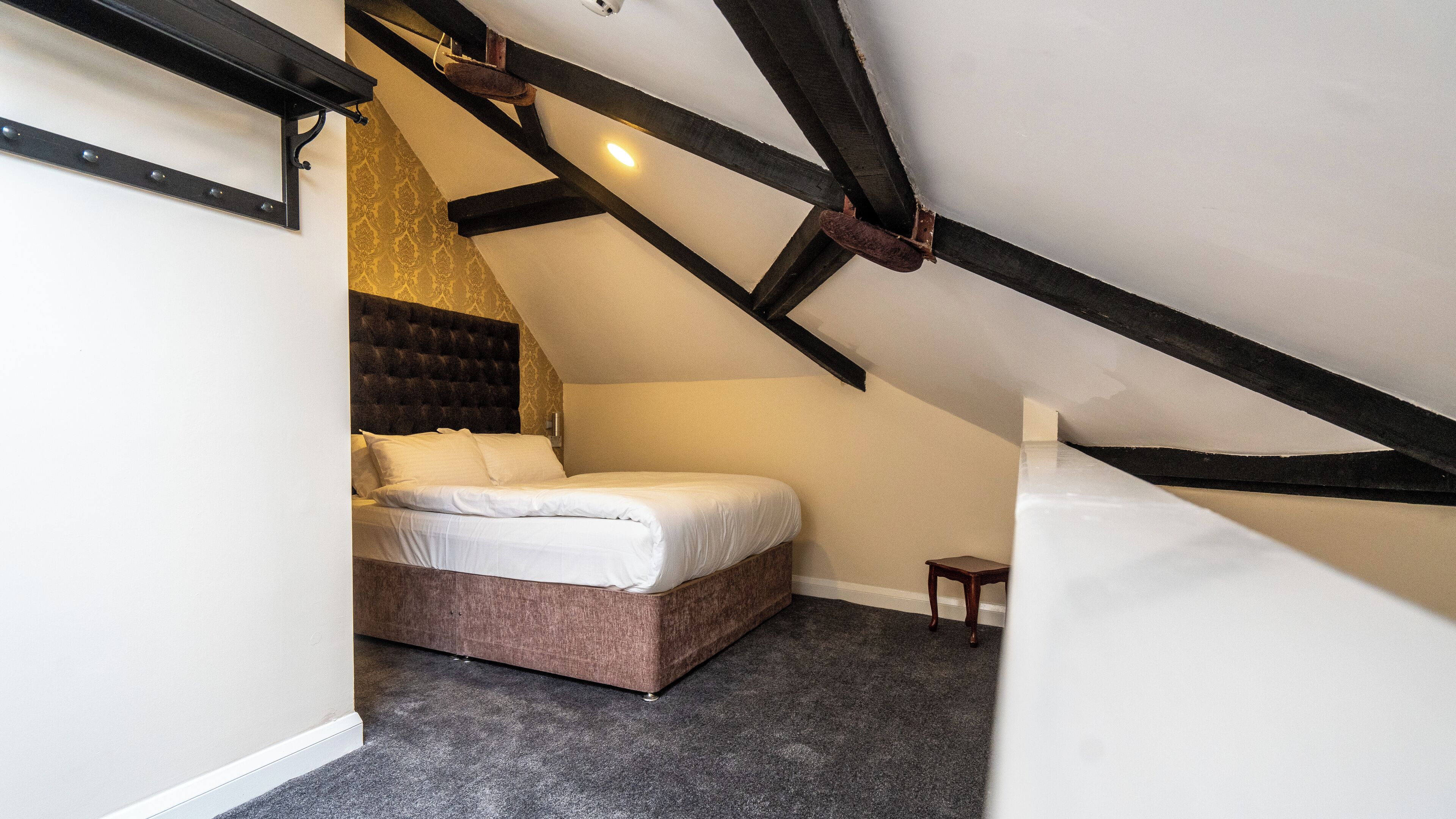Un dormitorio con techo inclinado, una cama con ropa de cama blanca, una mesita de noche de madera y una pared con estampado.