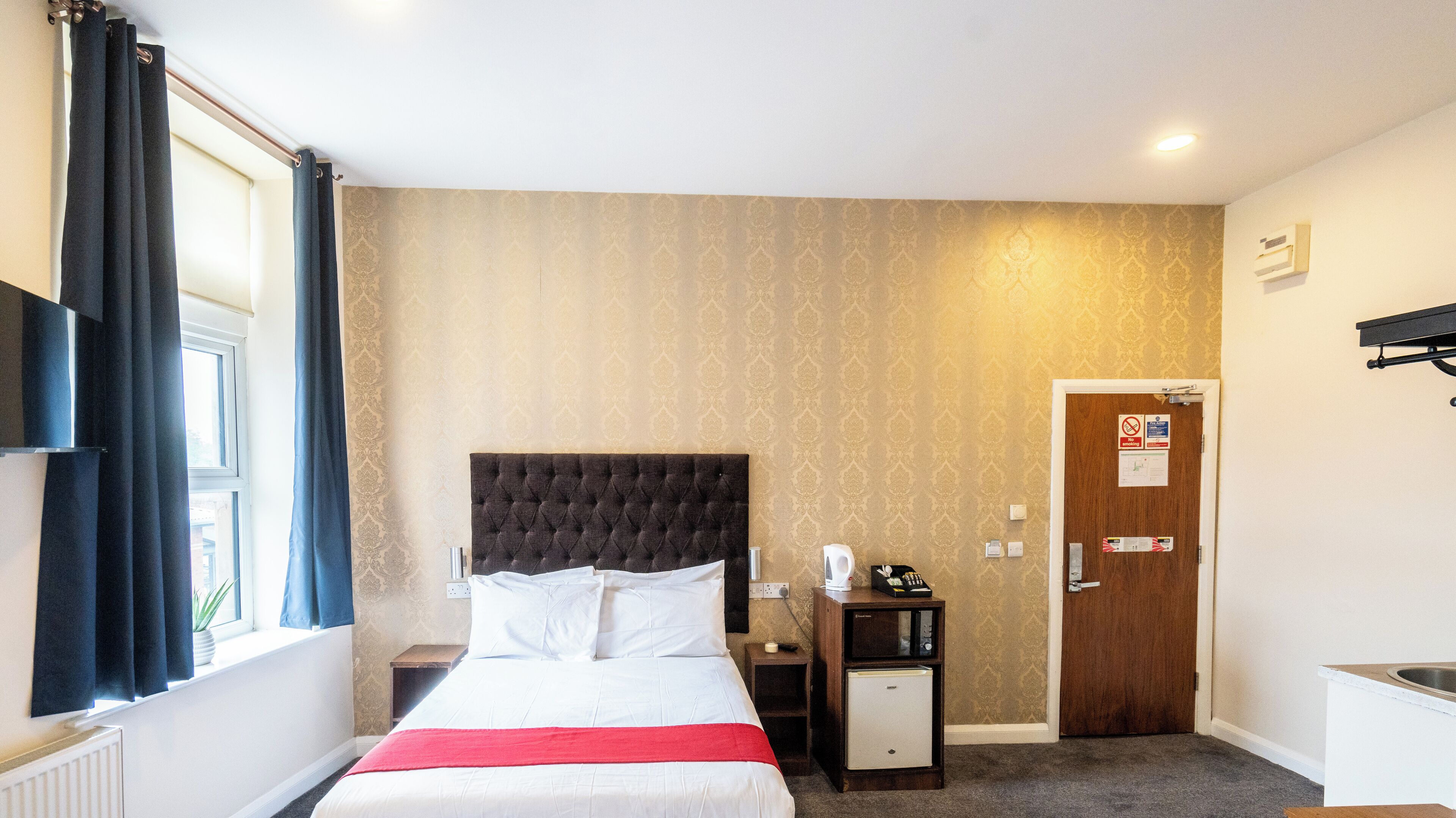 Une chambre d’hôtel avec un grand lit, une petite table de chevet, une commode, un bureau avec un lavabo et une porte avec une pancarte.