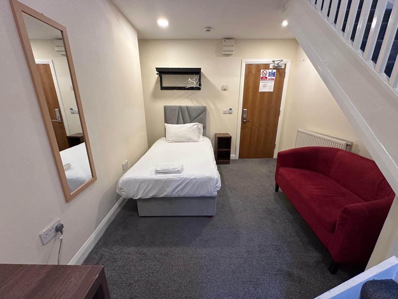 A small, neatly arranged room with a bed, a red sofa, a mirror, and a door with a sign.