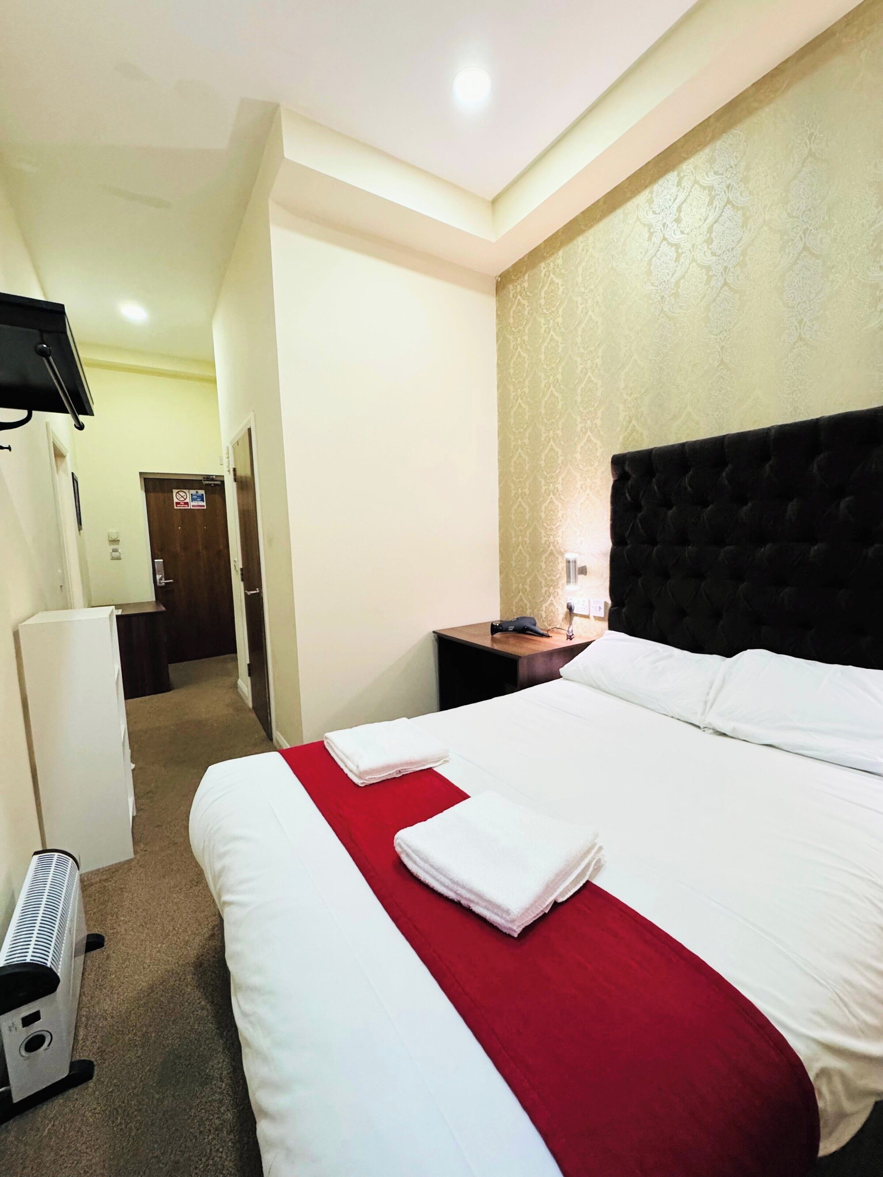 A hotel room with a large bed, a red throw blanket, a flat-screen TV mounted on the wall, and a desk with a lamp.
