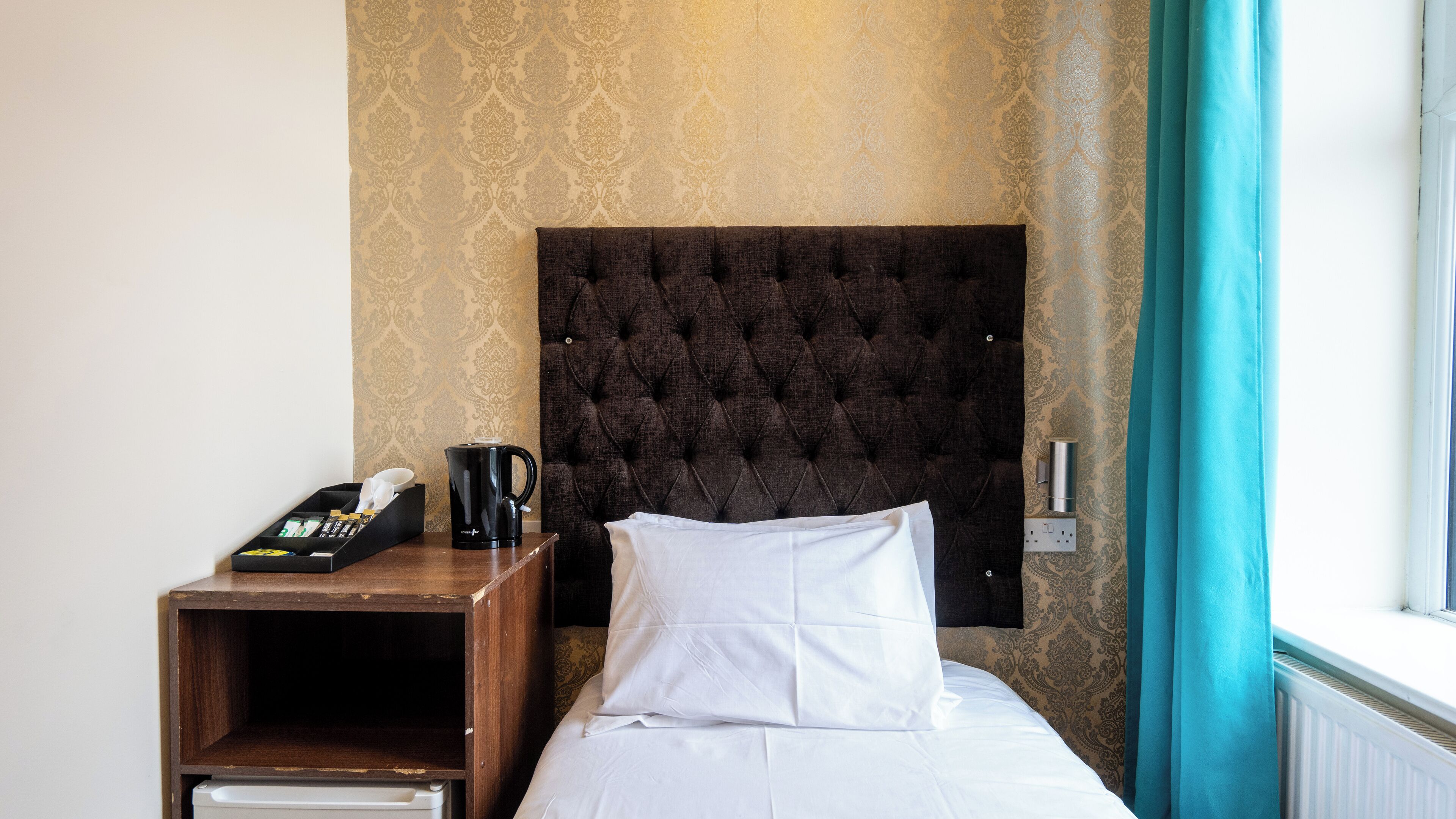 A hotel room with a tufted headboard, a bedside table with a coffee maker, and a window with curtains.