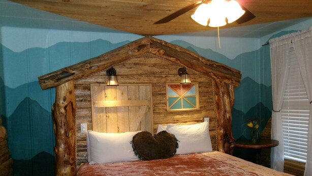A rustic bedroom with a wooden headboard, a bed with white and brown bedding, a bedside table with a vase, and a wall-mounted light fixture. - Hotel Eastin (Kremmling)