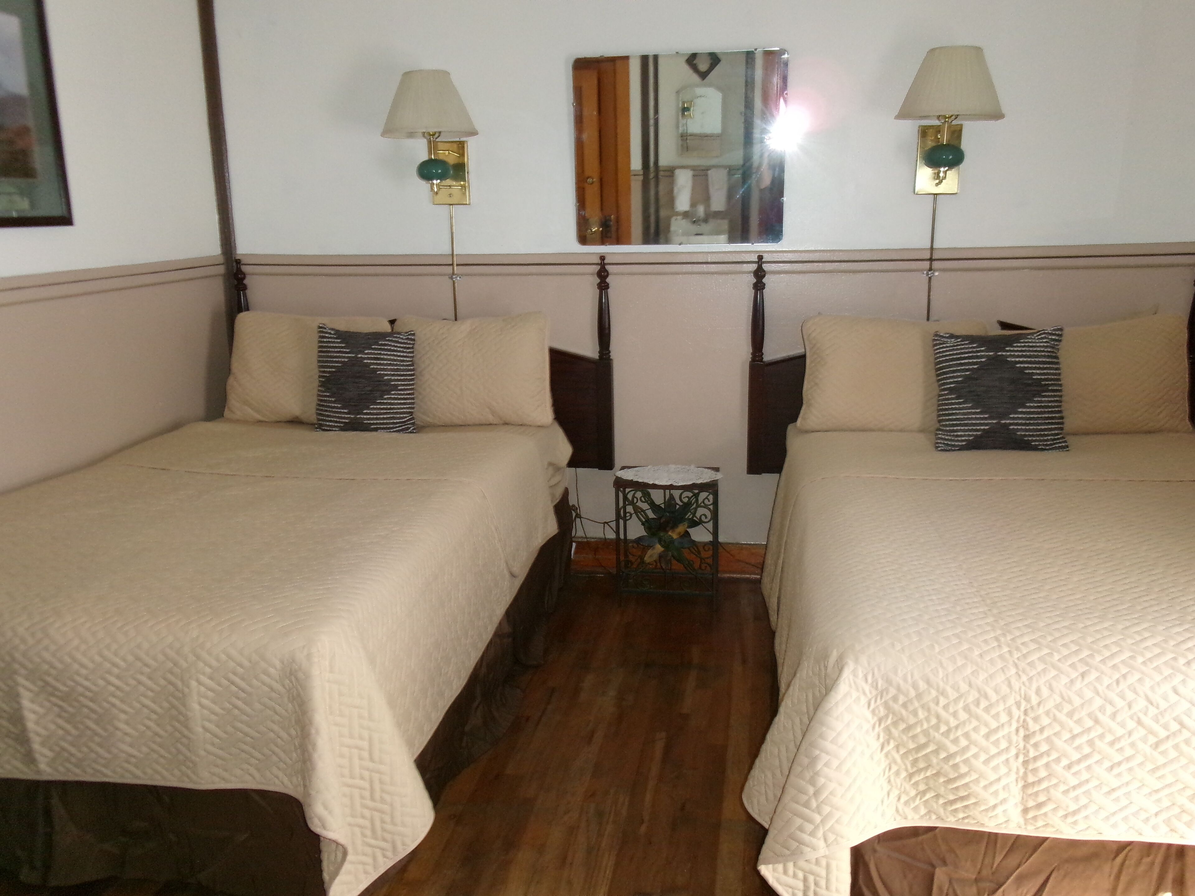Quarto com duas camas, cada uma com travesseiro e mesa de cabeceira. Há luminárias fixadas na parede e um espelho sobre as camas.