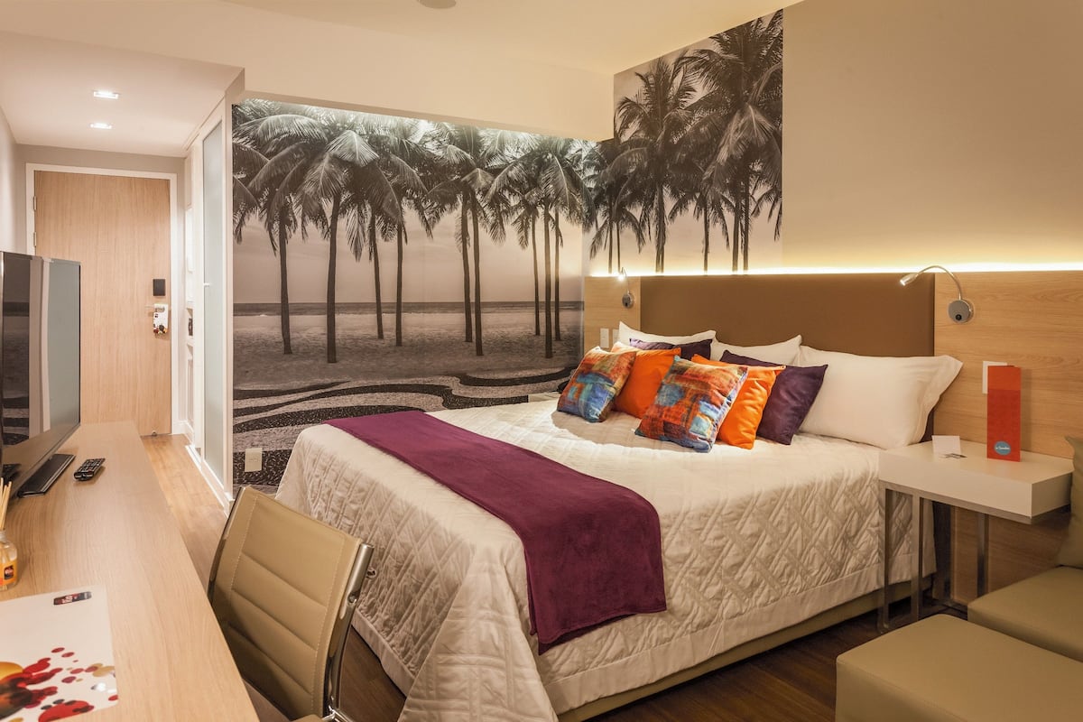 A hotel room with a bed, a desk with a computer, a chair, a TV, and a wall mural of palm trees.