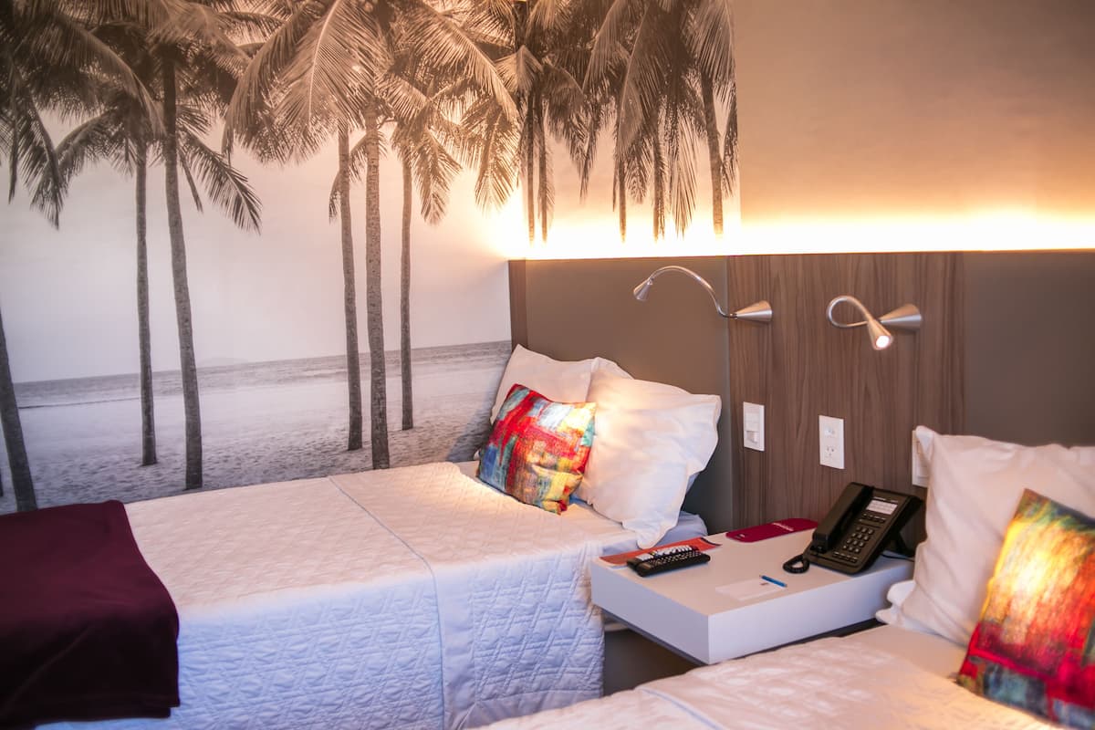 A hotel room with a bed, bedside table, lamp, and a view of the beach with palm trees.