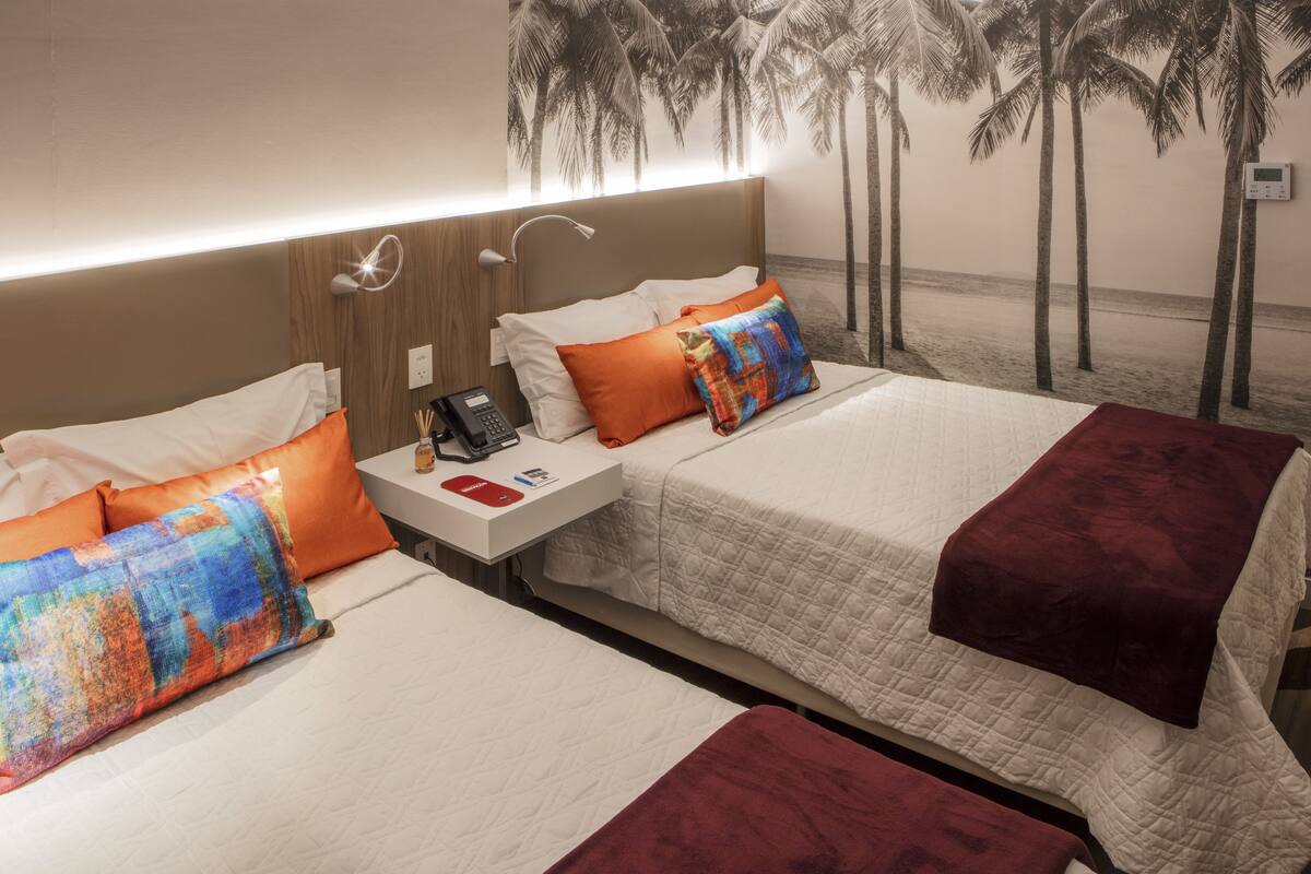 A hotel room with two beds, each with a white bedspread and orange pillows. There is a nightstand with a phone and a small table with a bottle and a box. The room has a headboard with a light fixture and a wall mural of palm trees.