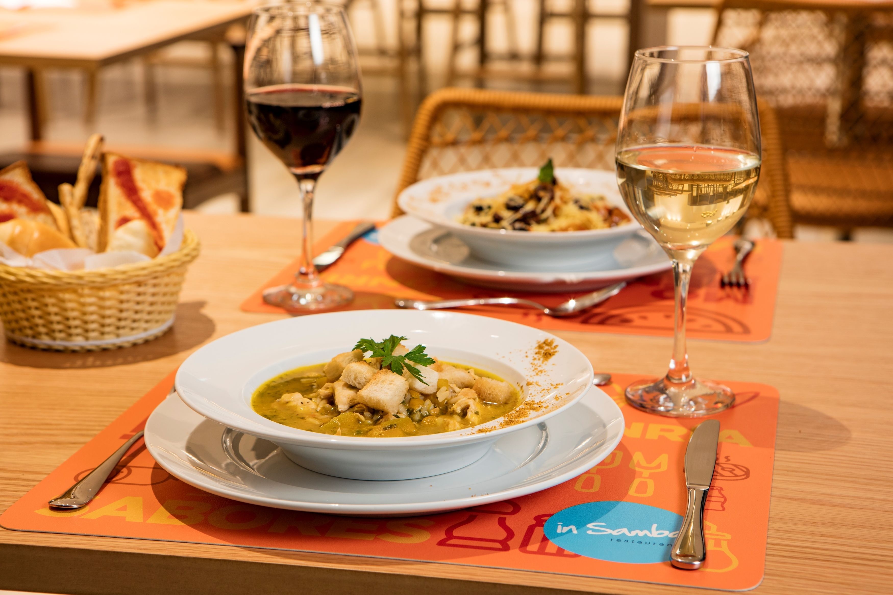 Una mesa de comedor con un tazón de sopa, dos copas de vino y una cesta de pan.