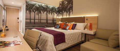 A hotel room with a bed, a desk, a chair, a sofa, and a wall mural of palm trees.