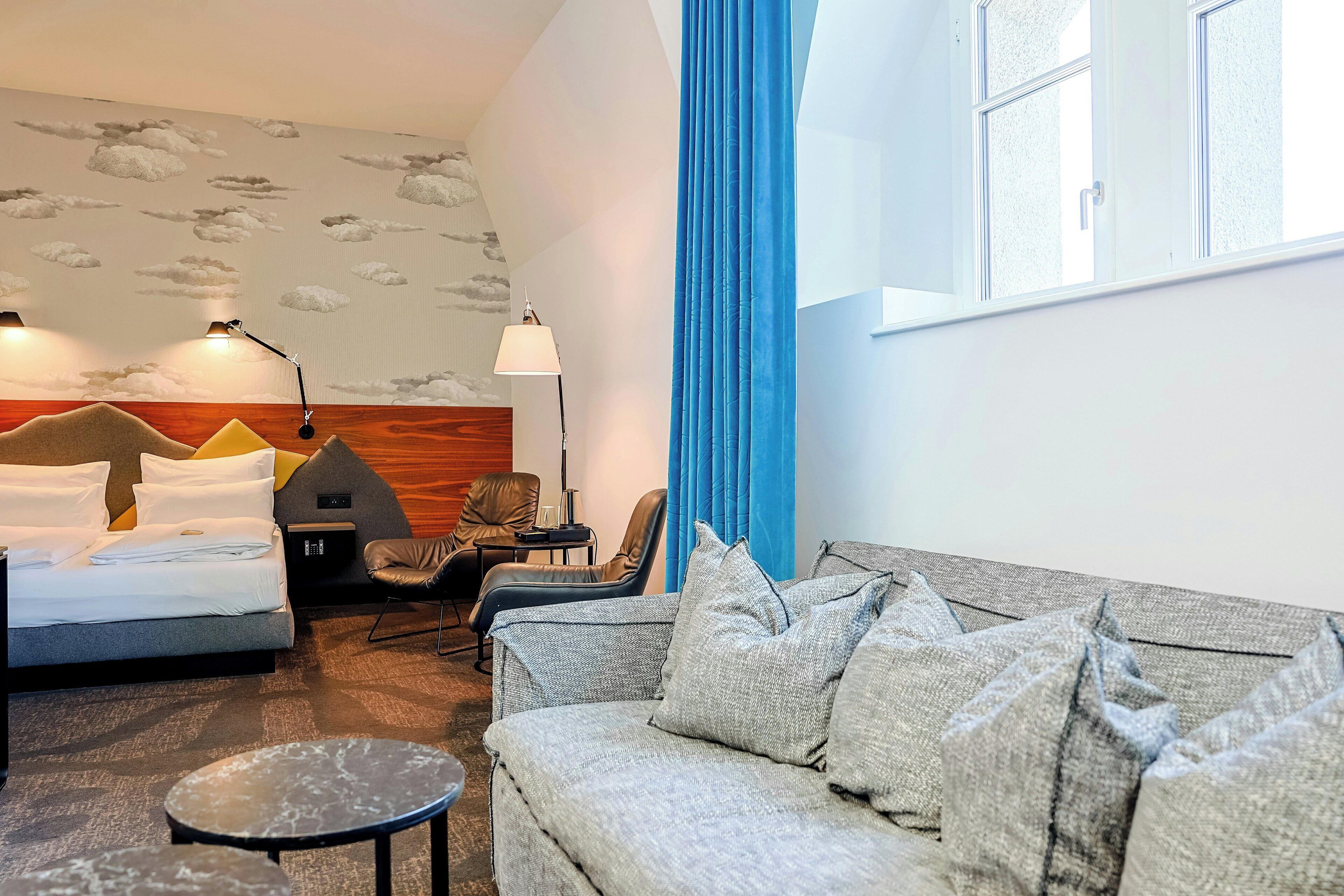 Habitación de hotel con cama, sofá, una mesita, silla, lámpara y ventana con cortinas.