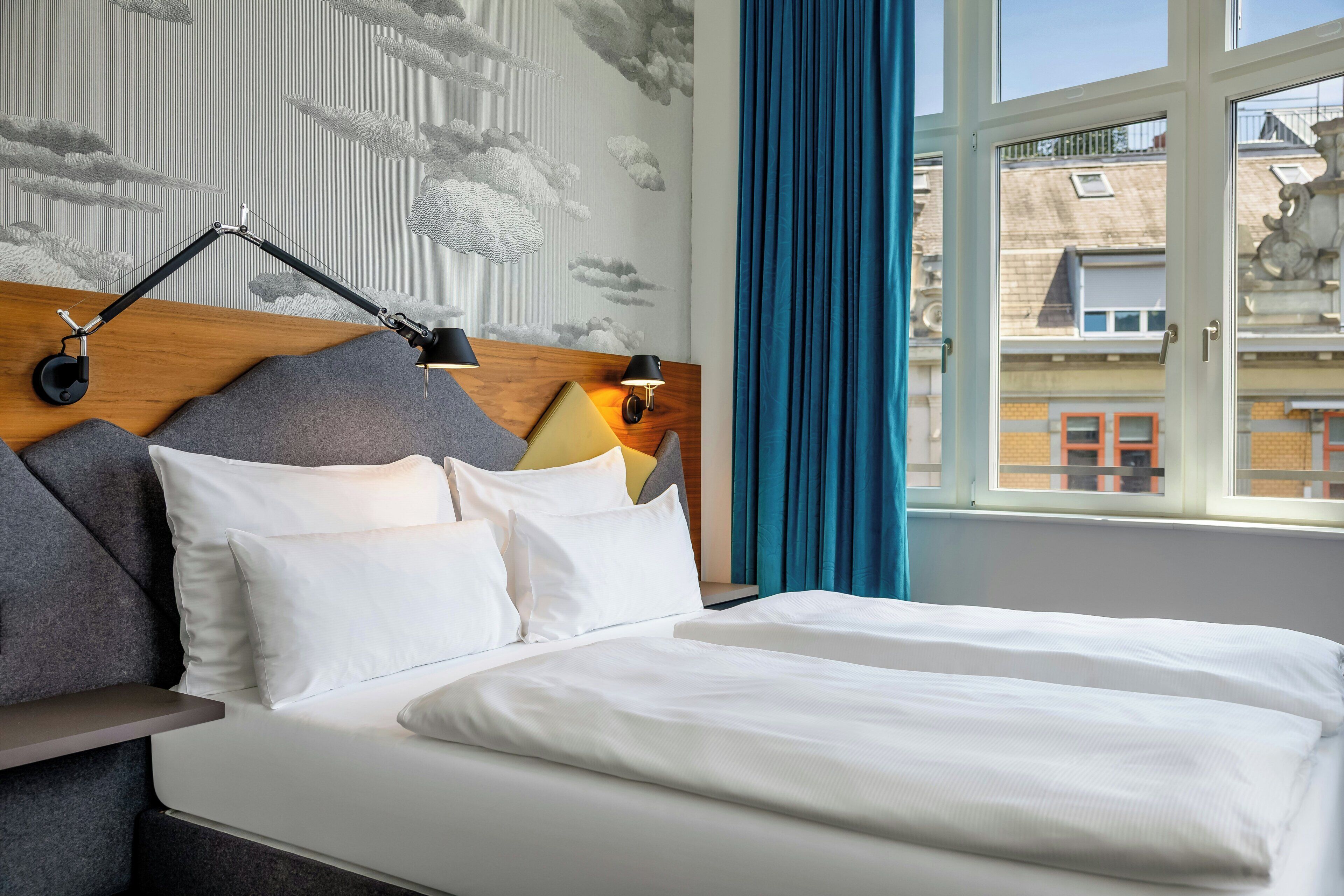 A hotel room with a bed, a headboard with a lamp, a window with curtains, and a view of buildings outside.