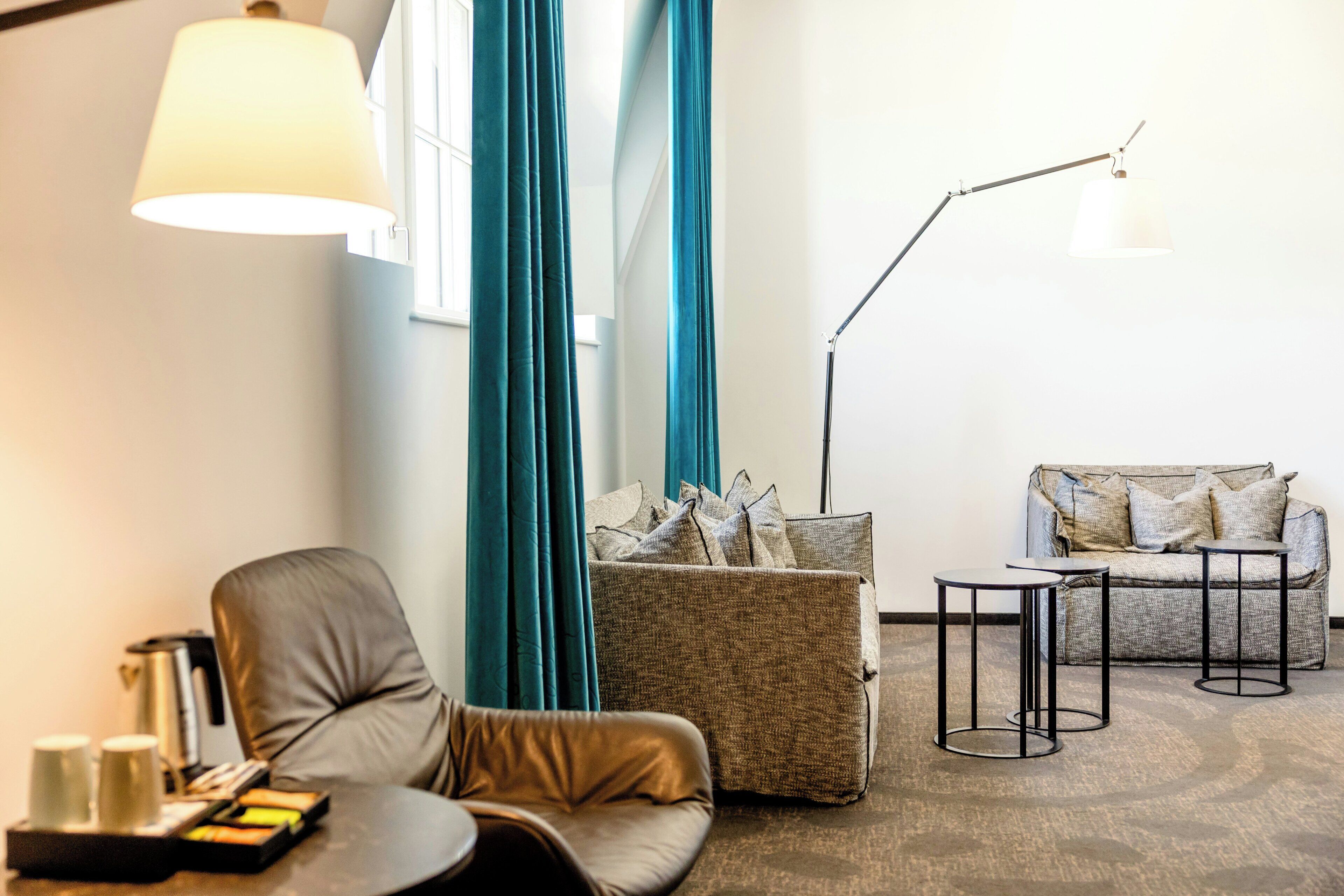 A modern living room with a leather chair, a sofa, two small tables, a floor lamp, and a coffee table with a tray of snacks.