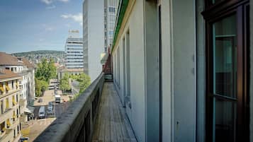 Un balcón estrecho con vistas a una calle, edificios, cielo despejado y colinas al fondo.