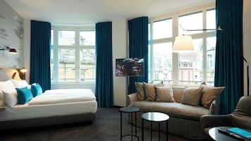 Habitación de hotel con cama, sofá, dos mesitas, televisión y ventanas grandes con cortinas.