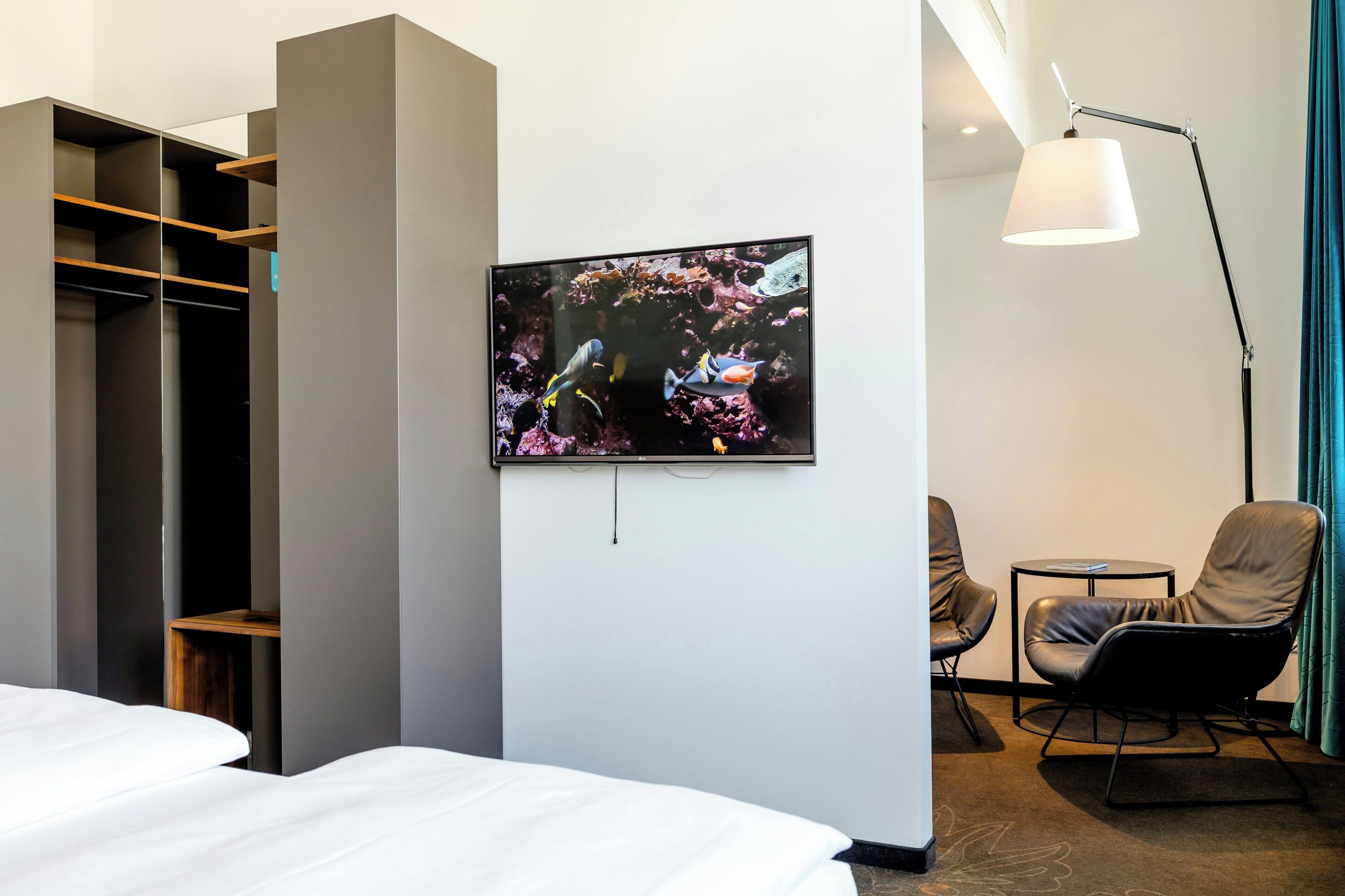 A hotel room with a bed, a TV mounted on the wall, a desk, a chair, and a wardrobe.
