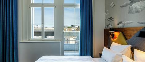 Habitación de hotel con un ventanal grande, una cama con ropa blanca y vistas a edificios.