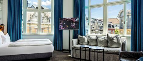 A hotel room with a bed, a sofa, a TV, and a view of the cityscape through large windows.