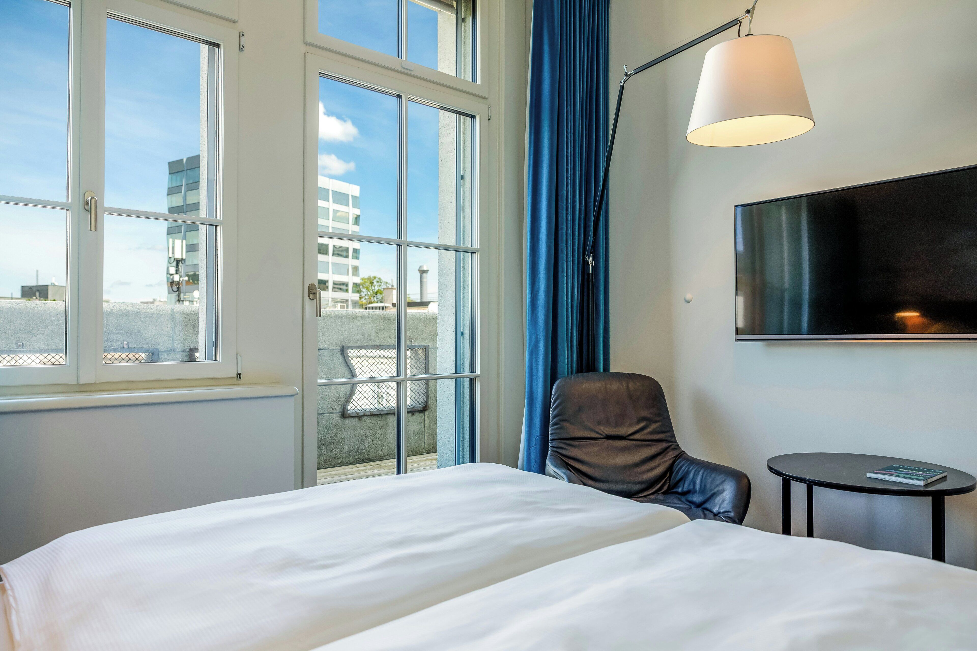 A hotel room with a bed, a chair, a small table, a TV, and a window with curtains.