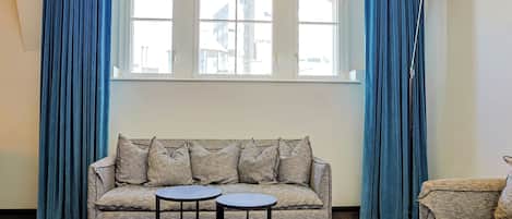Una habitación con sofá, dos mesitas, lámpara y ventana con cortinas.