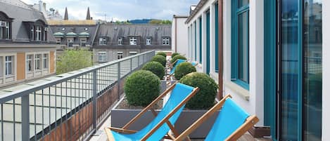 A balcony with wooden chairs and potted plants, overlooking a cityscape.