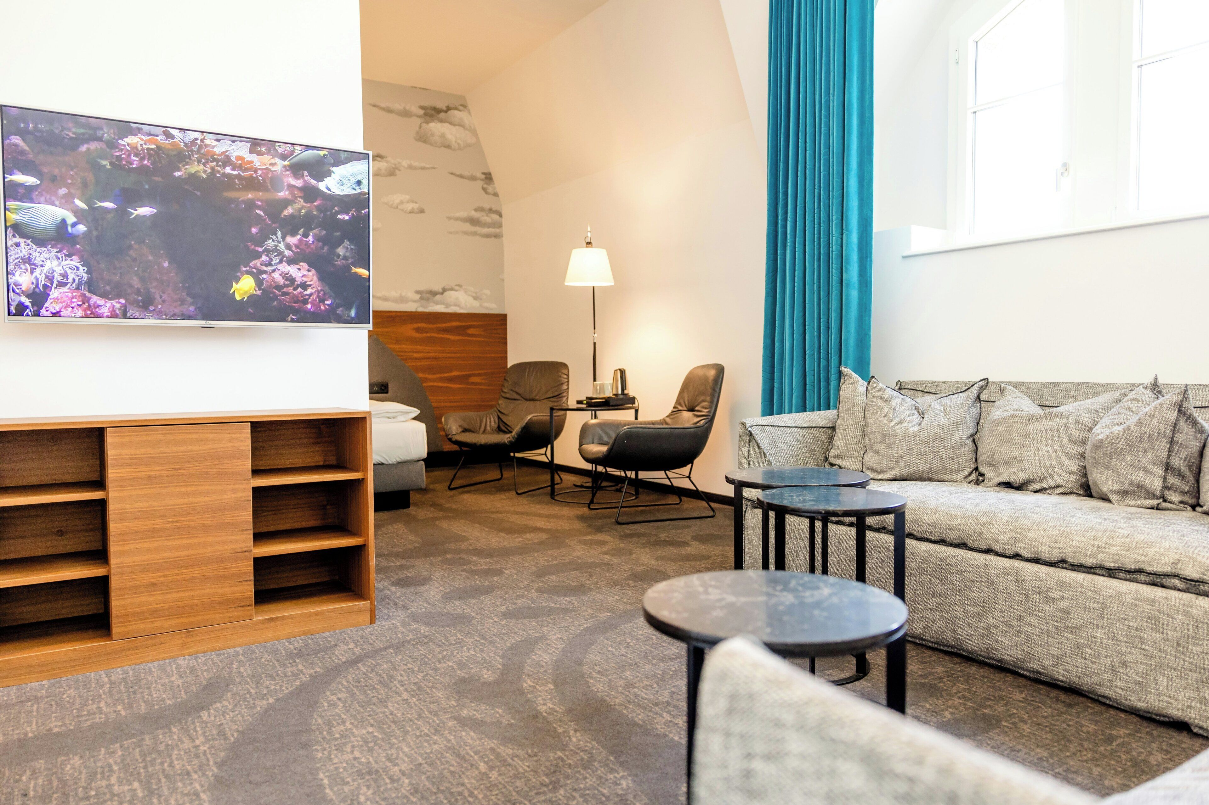A modern hotel room with a flat-screen TV, a sofa, two armchairs, a coffee table, and a lamp.