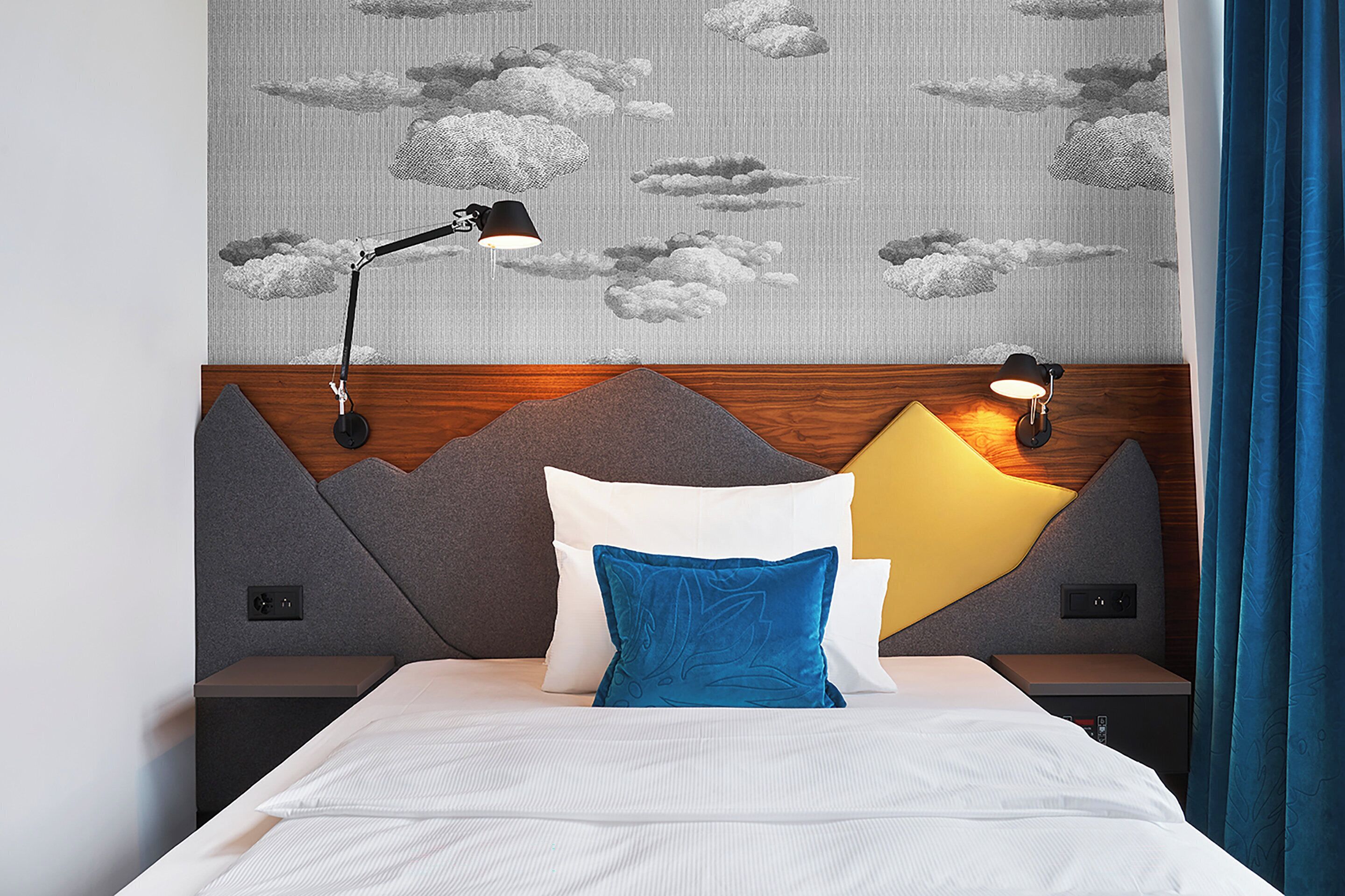 Ein ordentlich bezogenes Bett mit blau-weiß gestreiftem Kopfkissen, einem Holzkopfstück und einer Wand mit Wolkenmuster.
