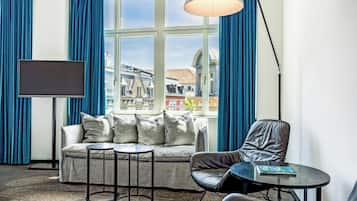 Ein modernes Zimmer mit einer Couch, zwei Beistelltischen, einem Sessel, einem Fernseher und einem Fenster mit Vorhängen.
